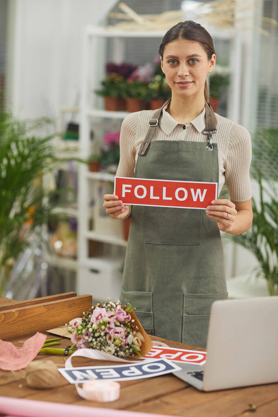 Vertical portrait of smiling young woman holding FOLLOW sign while standing in flower shop - Photo, Image