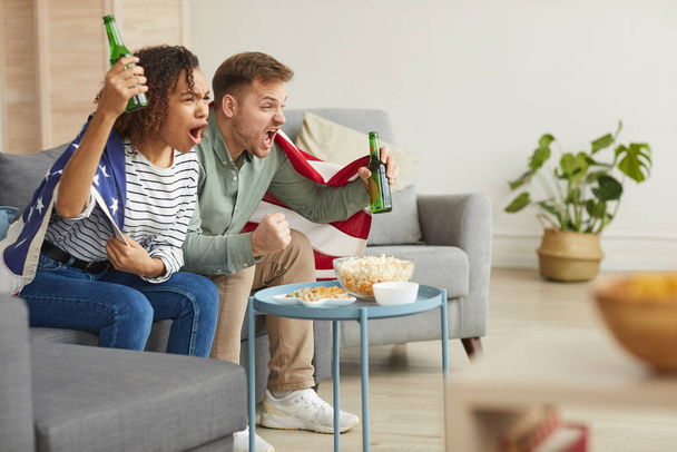 Side view portrait of young couple watching sports match on TV at home and cheering emotionally while wearing American flag, copy space - Photo, Image