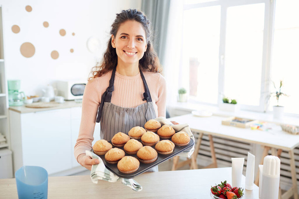 Moderne blanke vrouw houdt schimmel met hete cupcakes uit bakoven genomen kijken naar camera glimlachen - Foto, afbeelding
