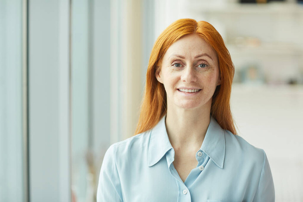 Head and shoulders portrait of red haired businesswoman smiling at camera while standing by window in modern white office, copy space - Photo, Image