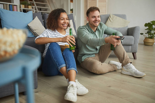 Retrato completo de casal misto moderno assistindo TV em casa e bebendo cerveja enquanto sentado no chão em apartamento acolhedor, espaço de cópia
 - Foto, Imagem