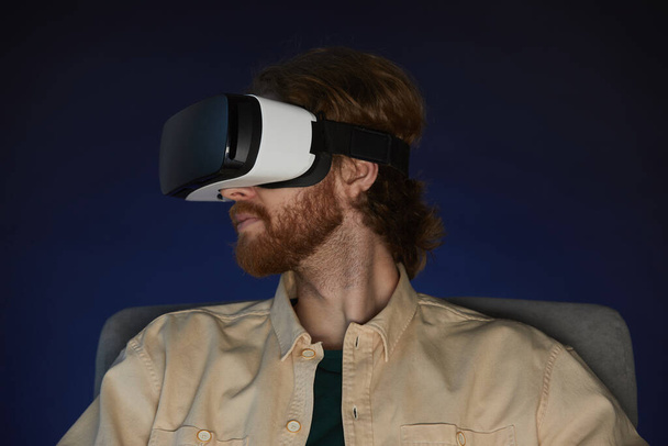 Πλάγια άποψη πορτρέτο του σύγχρονου γενειοφόρου άνδρα φορώντας εργαλεία εικονικής πραγματικότητας, ενώ απολαμβάνουν καθηλωτική videogame ή ταινία στο σκοτάδι - Φωτογραφία, εικόνα