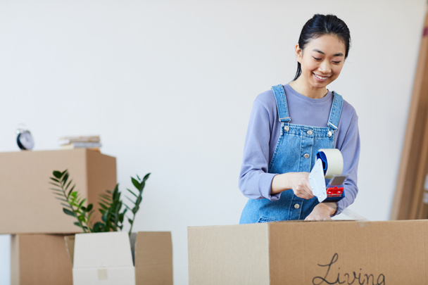 Waist up portrait of young Asian woman packing cardboard boxes with tape and smiling happily, excited for moving to new house, copy space - Photo, Image
