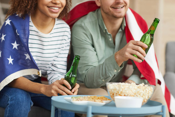Freigeschnittenes Porträt eines gemischten jungen Paares, das zu Hause fernsieht und Bier trinkt, während es die amerikanische Flagge trägt, Kopierraum - Foto, Bild