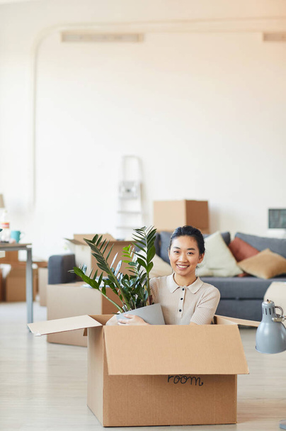 Retrato vertical de una joven asiática sentada en una caja y sosteniendo una planta mientras se muda a una nueva casa o apartamento, espacio para copiar
 - Foto, imagen