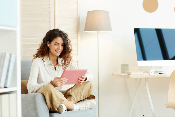 Mladá žena sedí uvolněně ve svém křesle a pracuje pomocí tabletu počítače a internetu - Fotografie, Obrázek