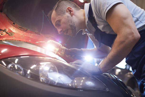 Close-up retrato de carro barbudo mecânico olhando sob o capô e usando lâmpada de lanterna durante a inspeção do veículo na garagem loja, espaço de cópia - Foto, Imagem