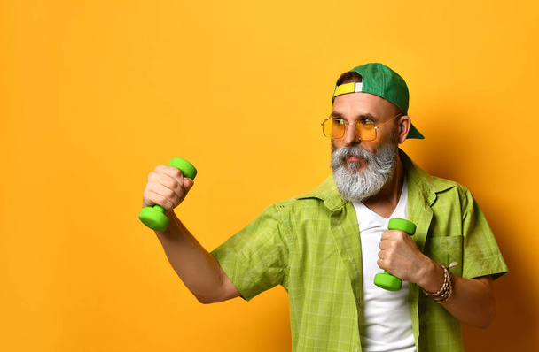 Grandpa in green cap and shirt, white t-shirt, bracelet. He smiling, doing exercises with dumbbells, posing on orange background - Фото, изображение