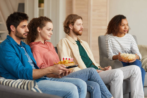 Multi-etnische groep vrienden die samen tv kijken terwijl ze thuis op een gezellige bank zitten en genieten van snacks, kopieer ruimte - Foto, afbeelding
