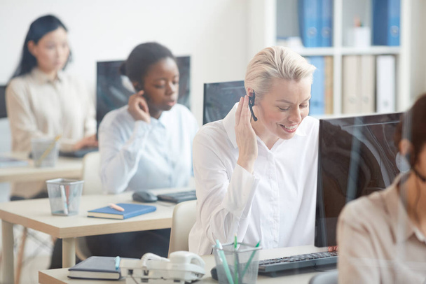 Portrait of female hotline operators sitting in row at desks , focus on smiling young woman talking to customer via headset, copy space - Photo, Image