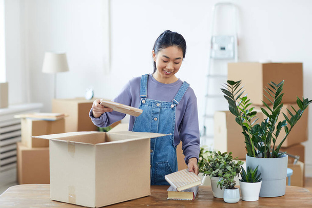 Waist up portrait of young Asian woman packing or unpacking cardboard box and smiling happily while moving into new home, copy space - Fotoğraf, Görsel