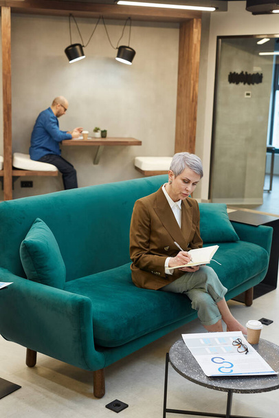Full length πορτρέτο της σύγχρονης ώριμης επιχειρηματία που γράφει στο βιβλίο σημείωμα, ενώ κάθεται στον καναπέ στο εσωτερικό του γραφείου σχεδιαστής, αντίγραφο χώρου - Φωτογραφία, εικόνα