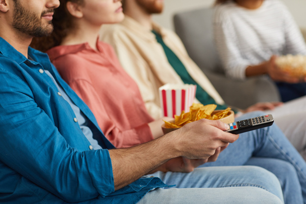 Close up of multi-ethnic group of friends watching TV together while sitting on cozy sofa at home and enjoying snacks, copy space - Фото, изображение