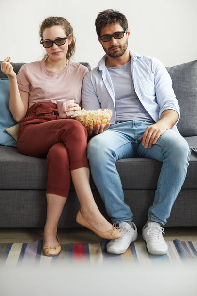 Κάθετη πλήρους μήκους πορτρέτο του σύγχρονου ζευγαριού βλέποντας τηλεόραση μαζί, ενώ κάθεται στον καναπέ και φορώντας γυαλιά ηλίου στο σπίτι απολαμβάνοντας τεμπέλης χρόνο - Φωτογραφία, εικόνα