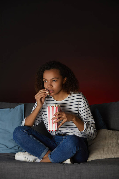 Vertikální celovečerní portrét mladé Afroameričanky, jak doma sleduje televizi a jí popcorn, zatímco sedí se zkříženýma nohama na gauči v tmavé místnosti, kopíruje prostor - Fotografie, Obrázek