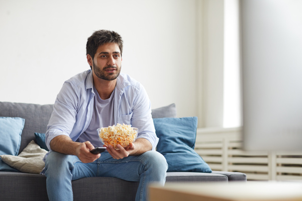 Retrato del hombre barbudo maduro viendo la televisión y sosteniendo un tazón de palomitas de maíz mientras está sentado en el sofá en casa, espacio para copiar
 - Foto, imagen