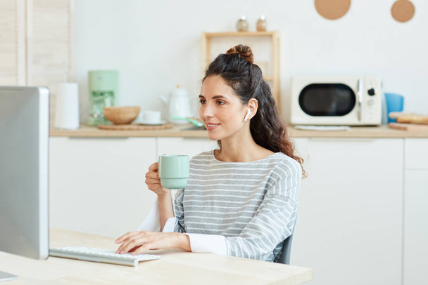 Горизонтальний середній портрет молодої жінки, яка використовує свій настільний комп'ютер і п'є ранкову каву вдома
 - Фото, зображення