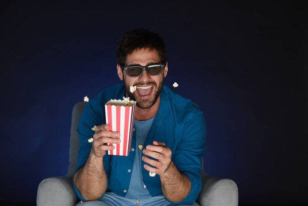 Portrait of joyful bearded man wearing 3D glasses eating popcorn while watching movie at home in dark, copy space - Foto, Imagem