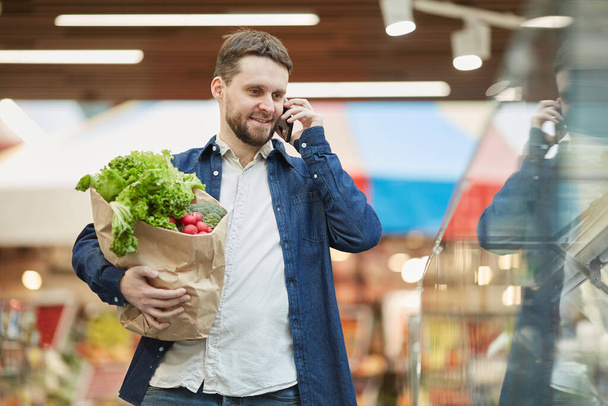Derékig portré mosolygós szakállas férfi hívja okostelefonnal, miközben élelmiszer-vásárlás a szupermarketben, másolás helyet - Fotó, kép