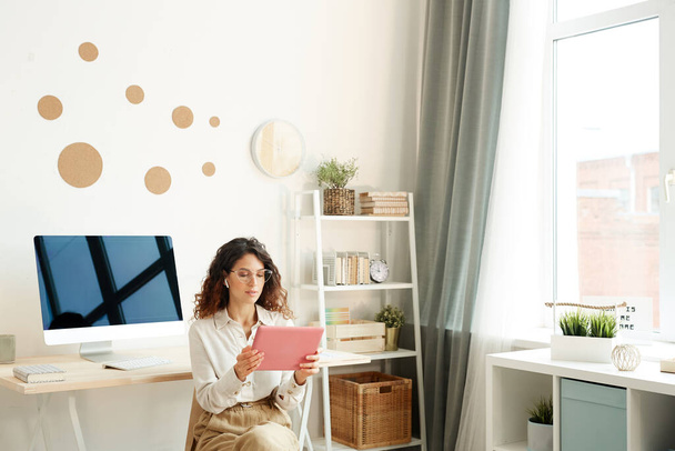 Junge Remote-Arbeiterin sitzt auf Stuhl in ihrem Zimmer und nimmt an Online-Treffen mit ihrem Tablet-Computer teil - Foto, Bild