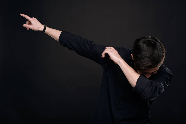 Νεαρός άνδρας με μαύρο πουκάμισο πέφτει με το κεφάλι σε απατεώνα του αγκώνα ενώ κάνει κίνηση dab σε απομονωμένο σκούρο φόντο - Φωτογραφία, εικόνα