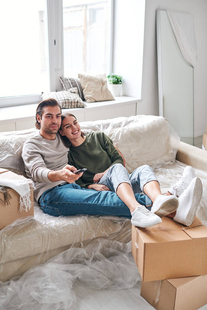 Glückliches junges erholsames Paar, das es sich auf der Couch gemütlich macht, während es mit den Beinen auf gepackten Kisten vor dem Fernseher sitzt, nachdem es in eine neue Wohnung gezogen ist - Foto, Bild