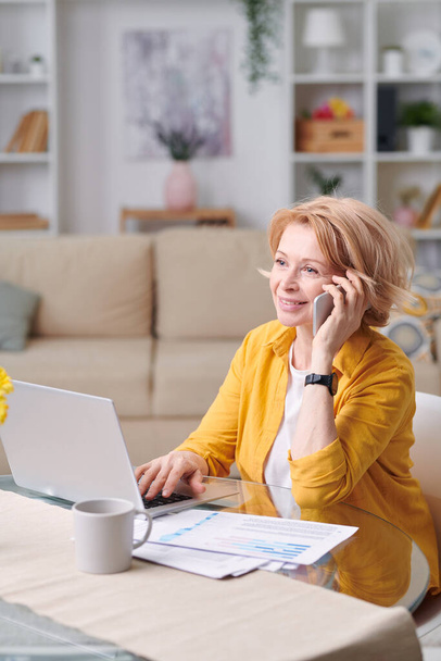 Volwassen vrolijke zakenvrouw met blond haar praten met iemand op de smartphone tijdens het werken op afstand thuis tijdens quarantaine - Foto, afbeelding
