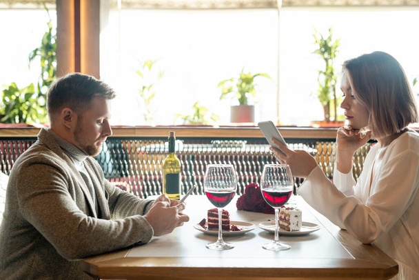 Młode współczesne daty przewijania w ich gadżety siedząc przy stole z ciastami i dwa kieliszki czerwonego wina w restauracji - Zdjęcie, obraz