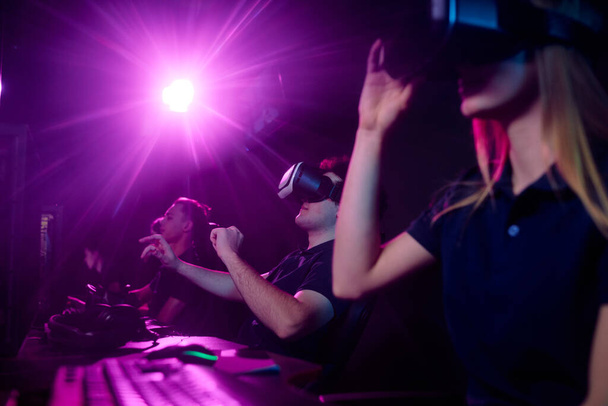 Groep van online gamers in virtual reality bril zitten voor krachtige computer en het spelen van video game in computer club met paars licht - Foto, afbeelding