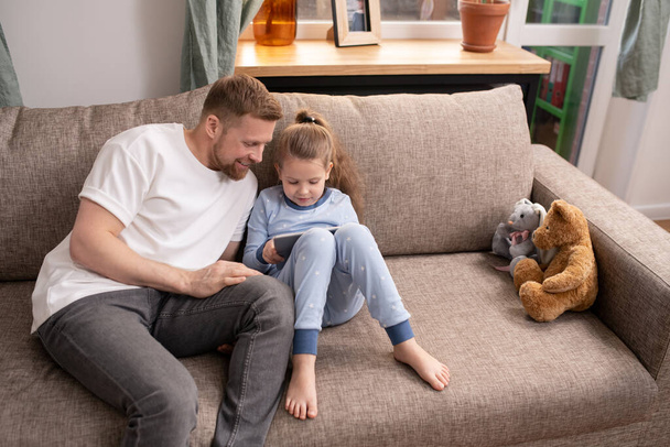 Nettes kleines Mädchen im blauen Pyjama und ihr glücklicher Vater in Freizeitbekleidung sitzen auf der Couch und schauen sich zu Hause Online-Filme im Touchpad an - Foto, Bild