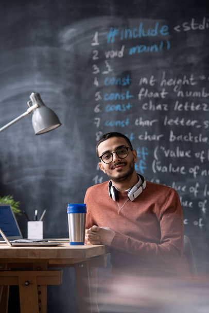 Портрет улыбающегося красивого молодого средневосточного веб-разработчика в очках, сидящего за столом с ноутбуком и термосом в офисе
 - Фото, изображение