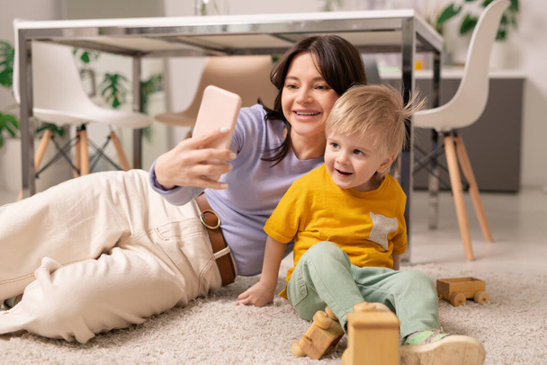 Onnellinen nuori äiti makaa matolla ja käyttää älypuhelinta poseeratessaan pienen pojan kanssa selfielle
 - Valokuva, kuva