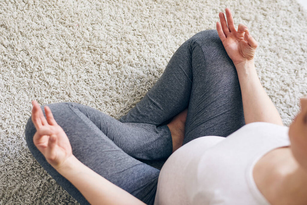 Vista superior de una joven embarazada en leggins sentada con las piernas cruzadas en la alfombra y cogida de la mano en mudra mientras medita en casa
 - Foto, imagen