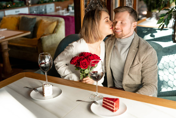 Νεαρή ευτυχισμένη γυναίκα με πολυτελές μπουκέτο κόκκινα τριαντάφυλλα φιλάει το αγόρι της στο μάγουλο, ενώ και οι δύο κάθονται δίπλα στο τραπέζι στο εστιατόριο - Φωτογραφία, εικόνα