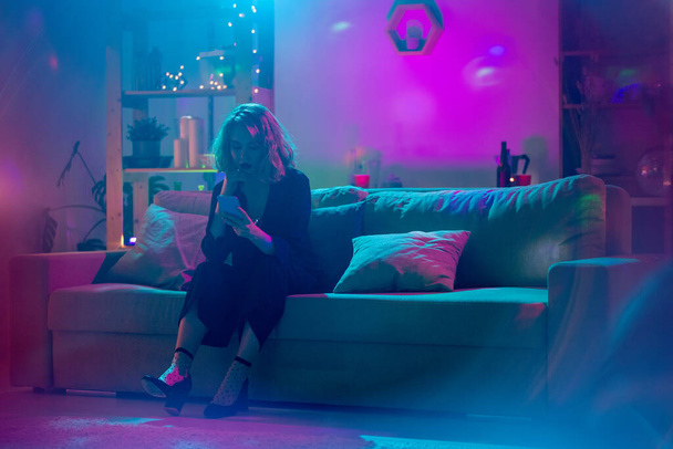 Fiatal szőke nő fekete elegáns öltöny ül a puha kanapén, és görgetés okostelefon a nappaliban világít rózsaszín és kék fény - Fotó, kép