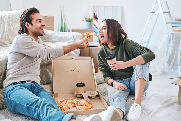 Glückliche junge Frau mit einem Glas Kaffee zum Mitnehmen, das den Mund öffnet, während ihr Mann ihr während des Mittagessens ein Stück Pizza auf dem Fußboden reicht - Foto, Bild