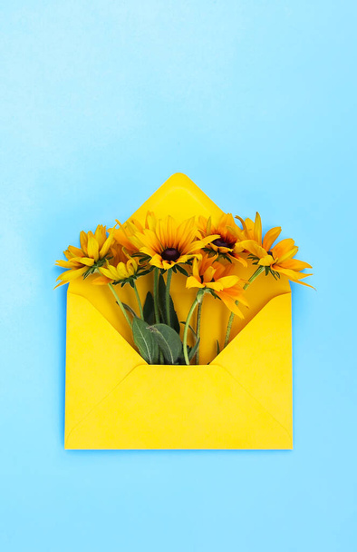 Yellow paper envelope with rudbeckia or black-eyed susan garden flowers on light blue background. Festive floral template. Greeting card design. Top view. Vertical shot. - Foto, Imagem