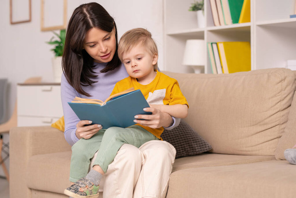 Mãe muito jovem lendo seu pequeno livro filho de contos de fadas enquanto segurando-o de joelhos durante o repouso em casa ou auto-isolamento
 - Foto, Imagem
