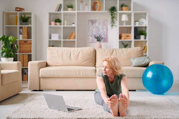Reife aktive Frau in Sportbekleidung sitzt auf dem Wohnzimmerboden vor dem Laptop und macht morgens Fitnessübungen - Foto, Bild