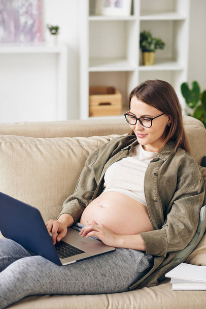Contenida joven embarazada con anteojos sentada en cómodo sofá y trabajando con laptop usando wi-fi en casa
 - Foto, imagen