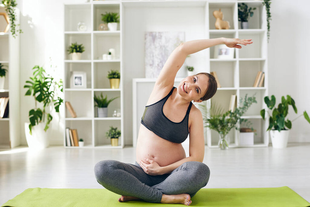 Muotokuva hymyilevä nuori raskaana oleva nainen urheiluasu istuu ristissä jalat ja taivutus syrjään kohotettu käsi harjoitellessaan joogaa kotona
 - Valokuva, kuva
