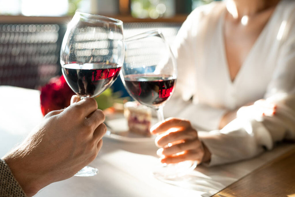 Hände eines jungen Mannes und seiner Freundin klirren mit Weingläsern über den Tisch, während sie ein romantisches Date und Mittagessen im luxuriösen Restaurant genießen - Foto, Bild