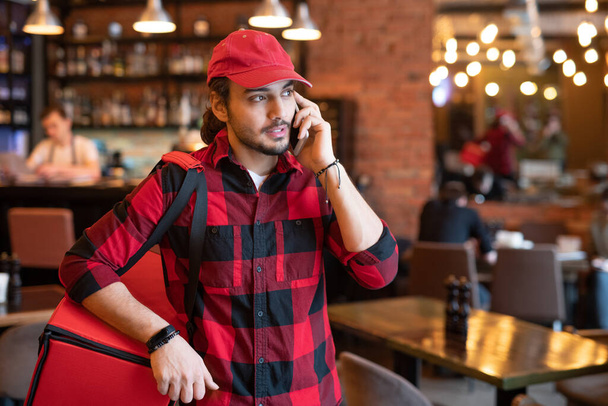 Zajęty kurier w odzieży roboczej rozmawia z jednym z klientów przez telefon komórkowy prosząc o adres i ostrzeżenie przed dostawą zamówionego jedzenia - Zdjęcie, obraz
