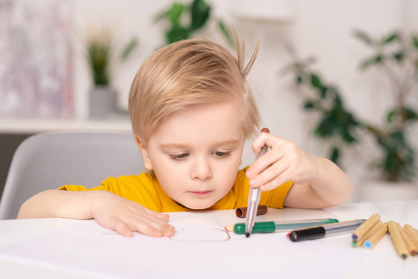 Masada oturup renkli keçeli kalemlerle kağıda resim çizen konsantre olmuş sarı saçlı çocuk. - Fotoğraf, Görsel
