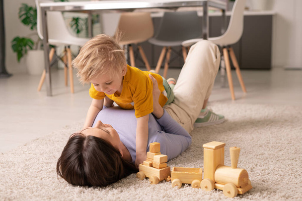 Bella giovane donna bruna sdraiata su un soffice tappeto sul pavimento e che gioca con il suo adorabile piccolo figlio mentre rimane a casa durante la quarantena
 - Foto, immagini