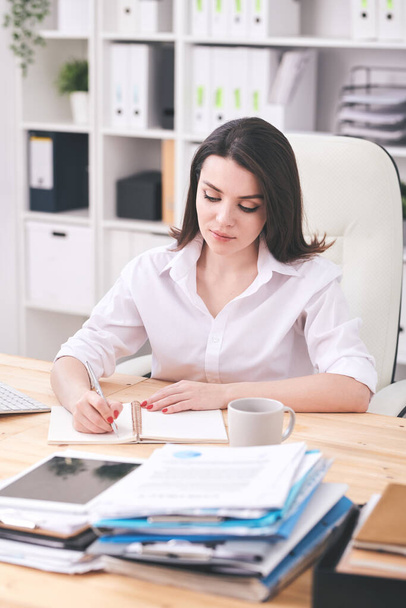 Jolie jeune femme sérieuse en chemise blanche assise dans un fauteuil par bureau et faisant un plan de travail dans un carnet sur fond d'étagères
 - Photo, image