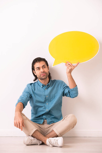 Pensive young man with stubble sitting with crossed legs on floor and holding speech bubble tag near head - Photo, Image