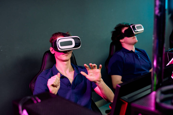 Curiosos jóvenes con las mismas camisetas sentados en el club oscuro e-sport y usando simuladores de realidad virtual mientras juegan videojuegos en red
 - Foto, imagen