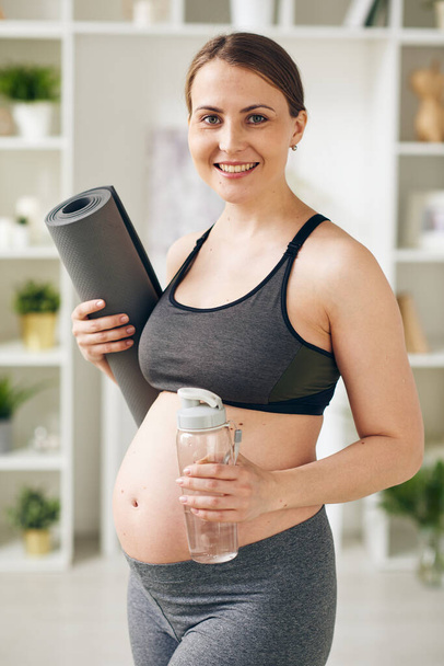 Портрет веселой молодой беременной женщины в серой спортивной одежде, стоящей с катанием коврика для йоги и фитнес-бутылки, она заботится о своем здоровье
 - Фото, изображение