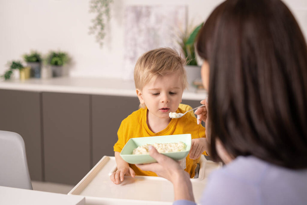 Schattige kleine jongen eten smakelijke zelfgemaakte pap terwijl zorgvuldige moeder houden lepel door zijn mond, terwijl het voeden van hem tijdens het ontbijt - Foto, afbeelding